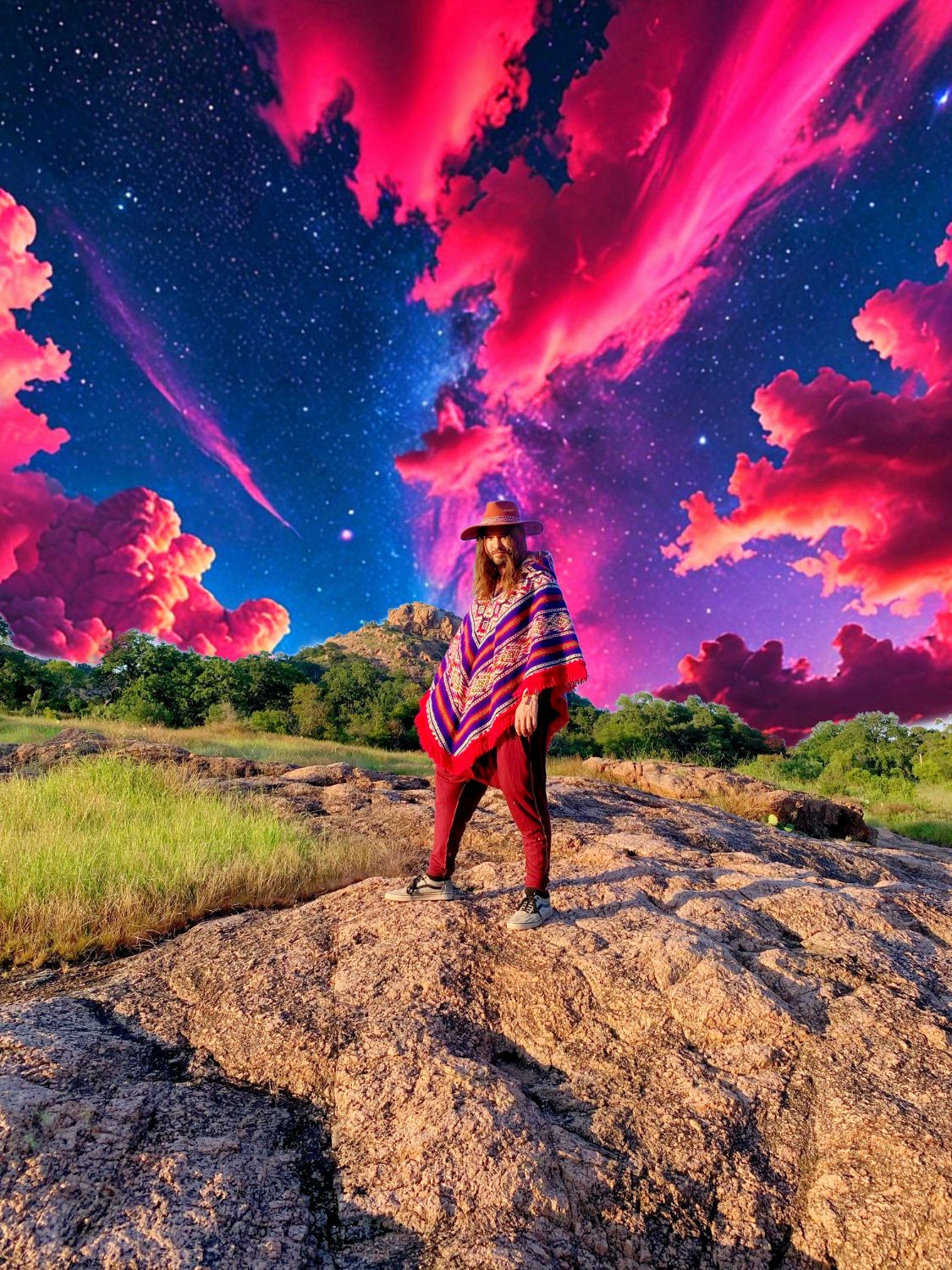 Daniel in a poncho and wide-brim hat standing on a granite outcrop under a pink and violet galaxy sky