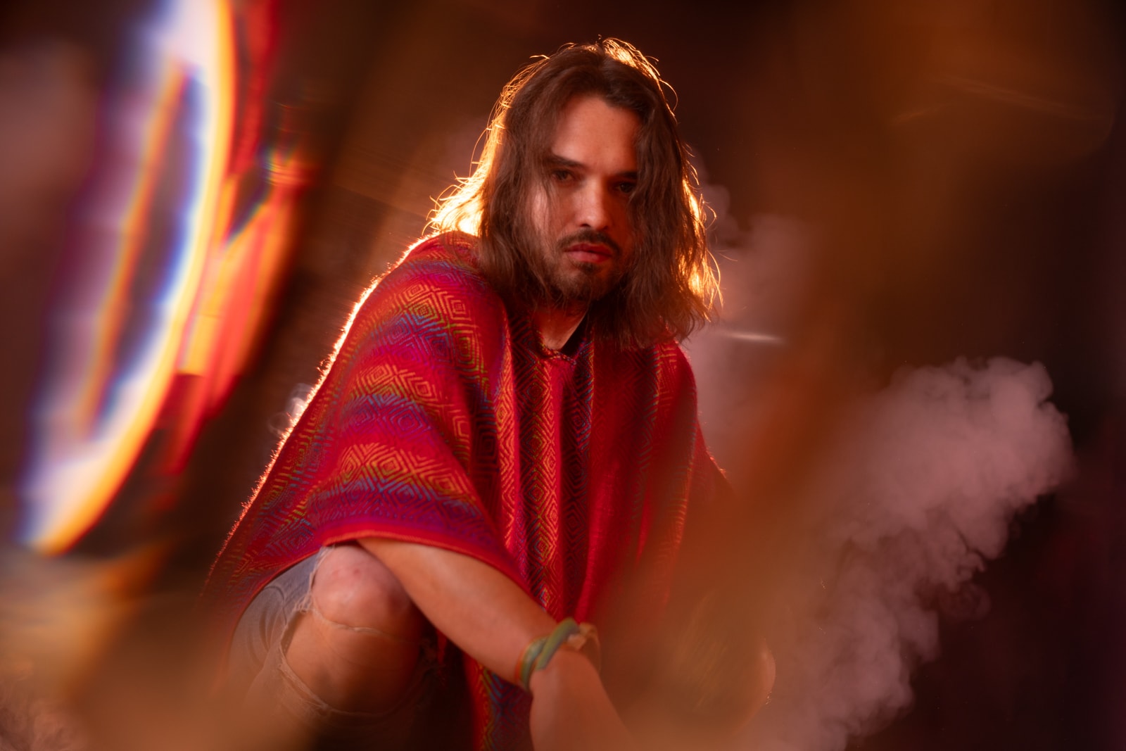Daniel crouched in a red woven poncho, backlit, with smoke and a ring of light around him