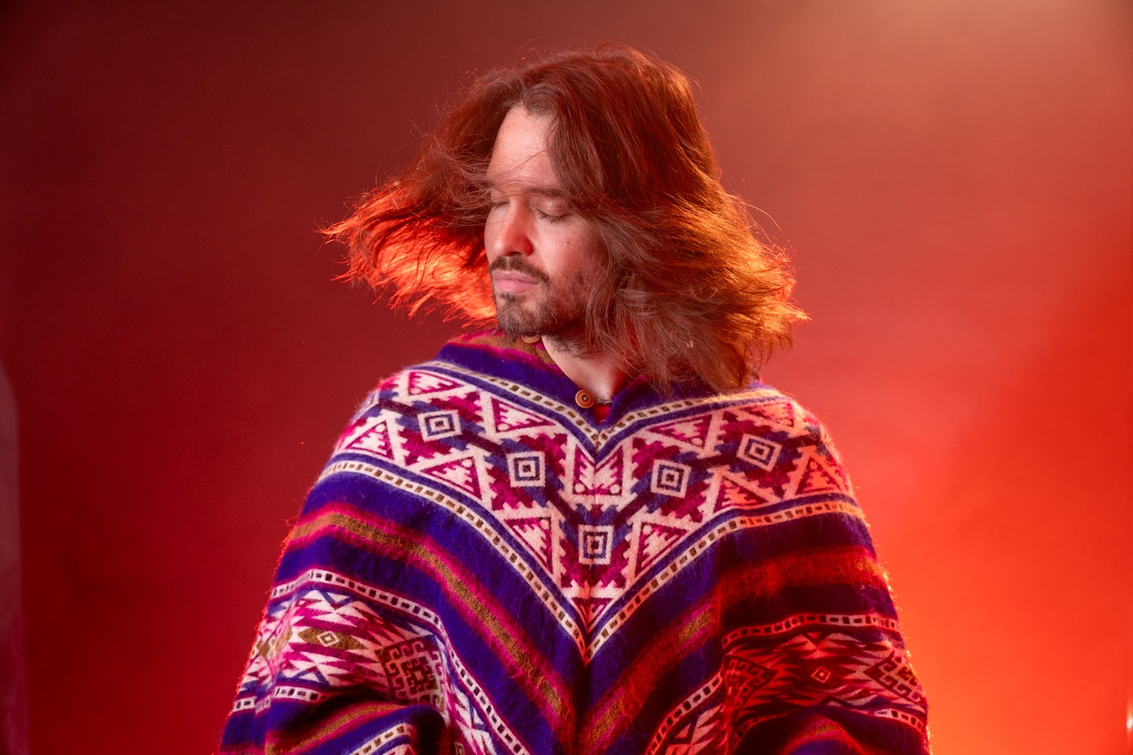 Portrait of Daniel in a patterned poncho, his hair in motion against a warm red backdrop